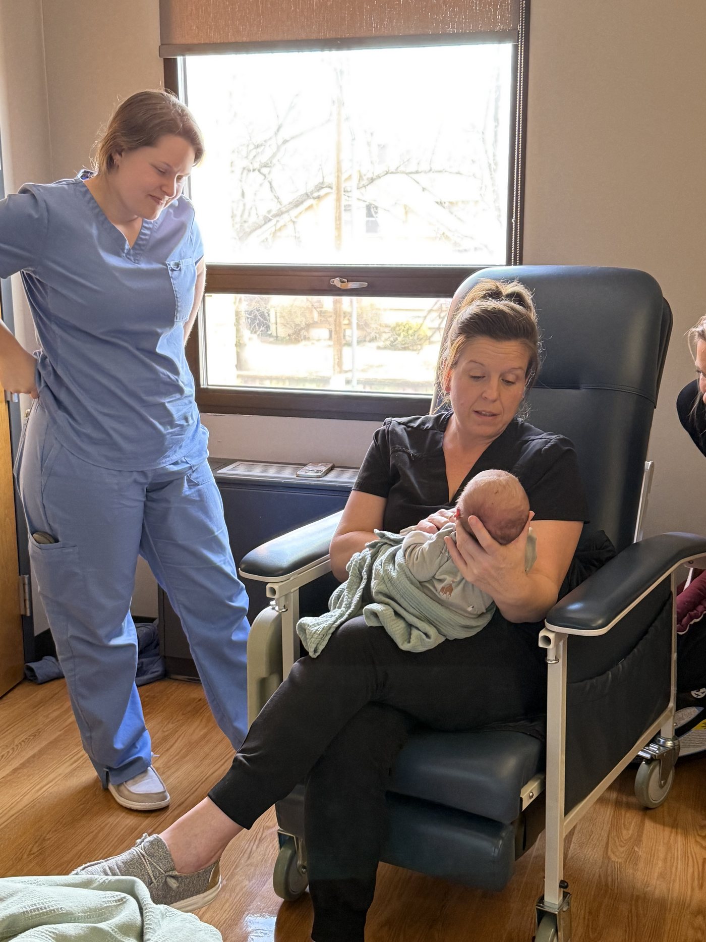 A mother resting in the birth suite recliner with her newborn while her midwife looks on